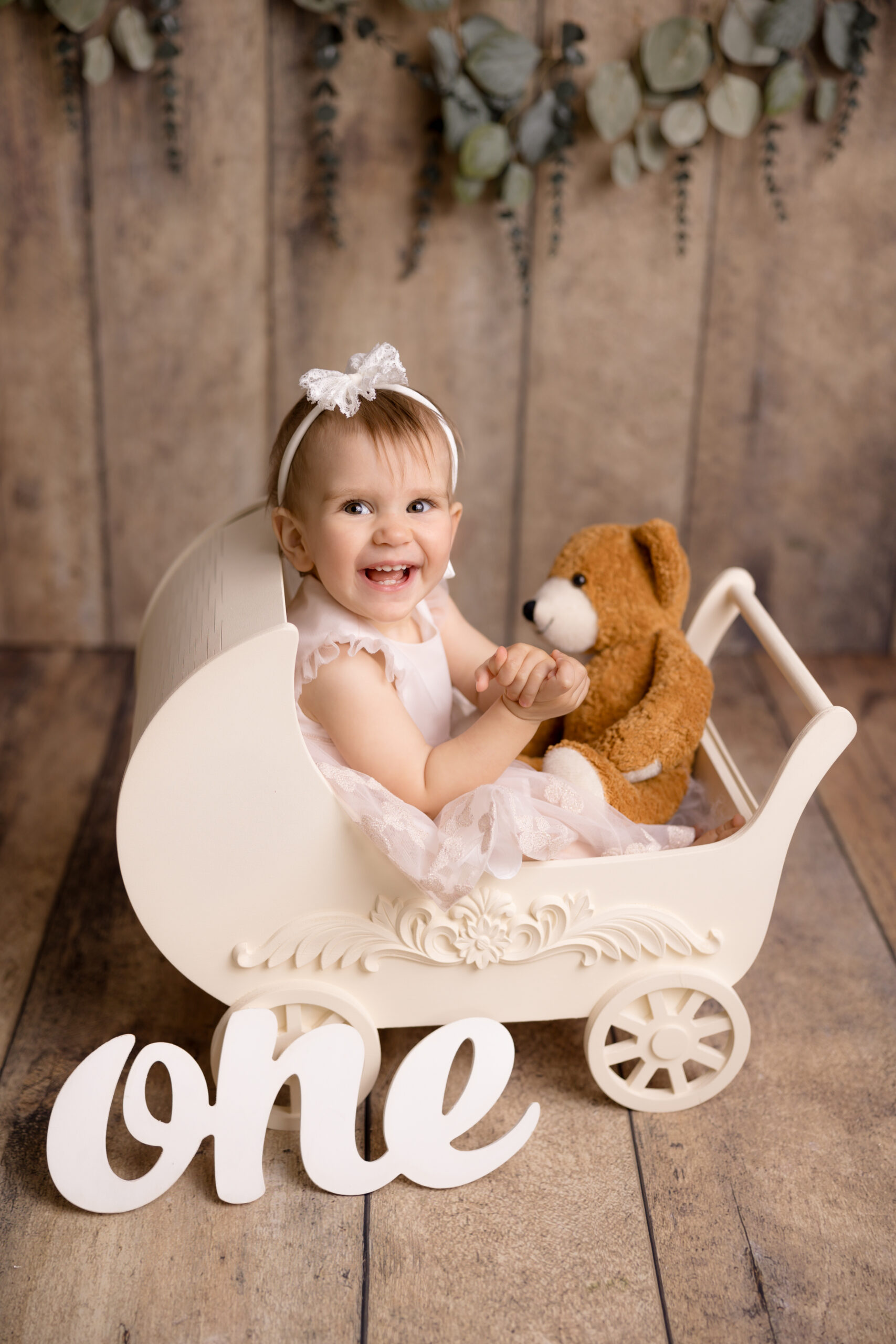cake smash first birthday photo session smiling baby girl is sitting in a wooden pram with teddies and letters ONE in Wimbledon photo studio South West London